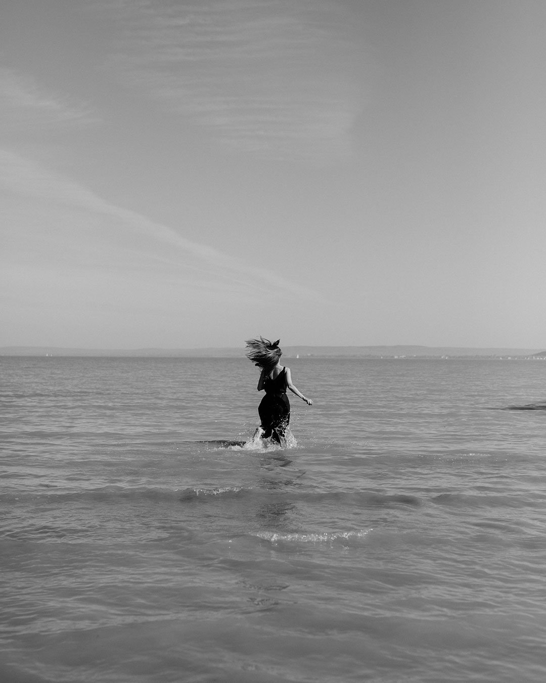 A woman running in the shallow water at the beach, with her hair flowing in the wind, in a black and white photo.