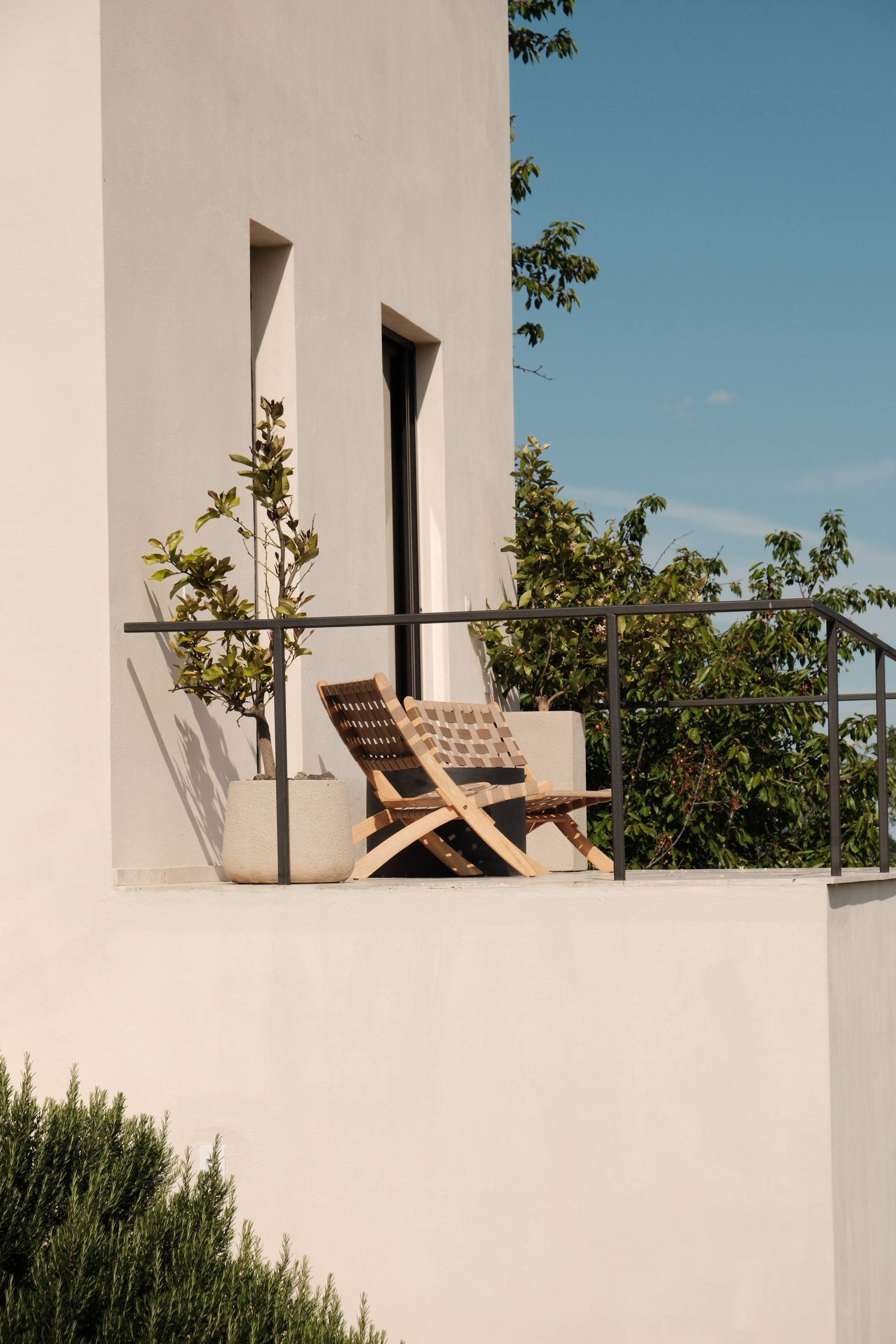 A balcony with a wooden chair and a small table, potted plants, and trees in the background under a clear blue sky.